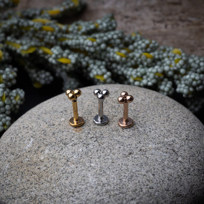 Close-up of three minimalist ball cluster labret studs in rose gold, silver, and gold finishes, displayed on a stone background.

