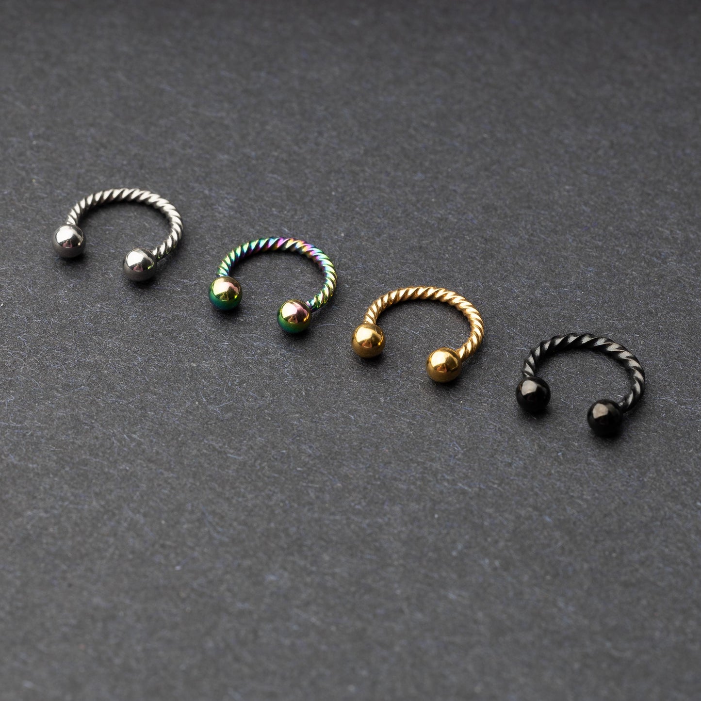 Twisted horseshoe barbells in silver, rainbow, gold, and black finishes displayed on a dark background, highlighting their bold and unique design.