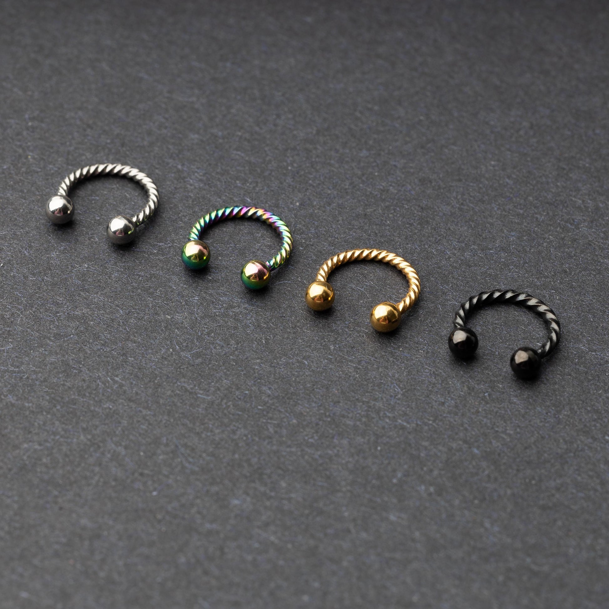 Twisted horseshoe barbells in silver, rainbow, gold, and black finishes displayed on a dark background, highlighting their bold and unique design.