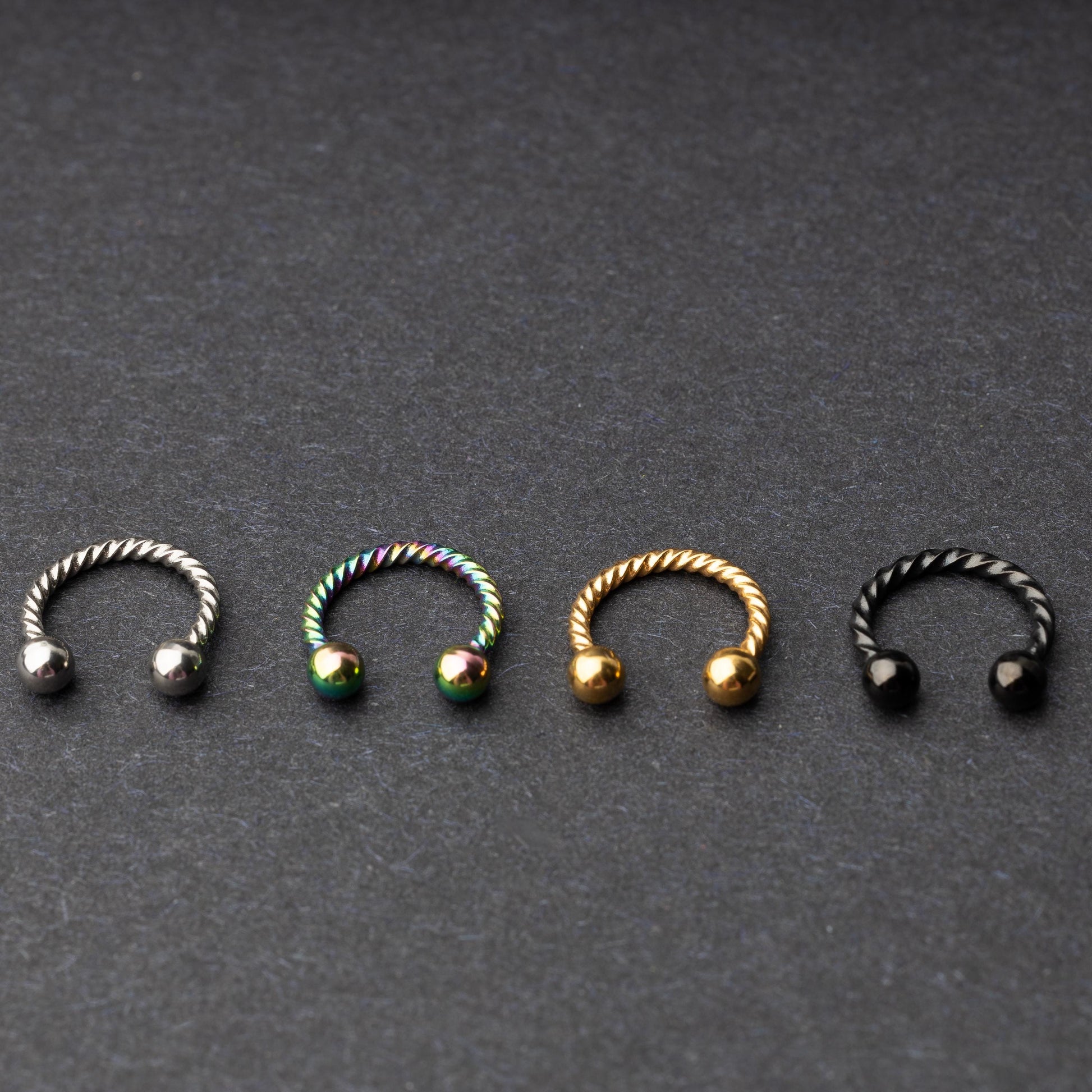 Twisted horseshoe barbells in silver, rainbow, gold, and black finishes displayed on a dark background, highlighting their bold and unique design.