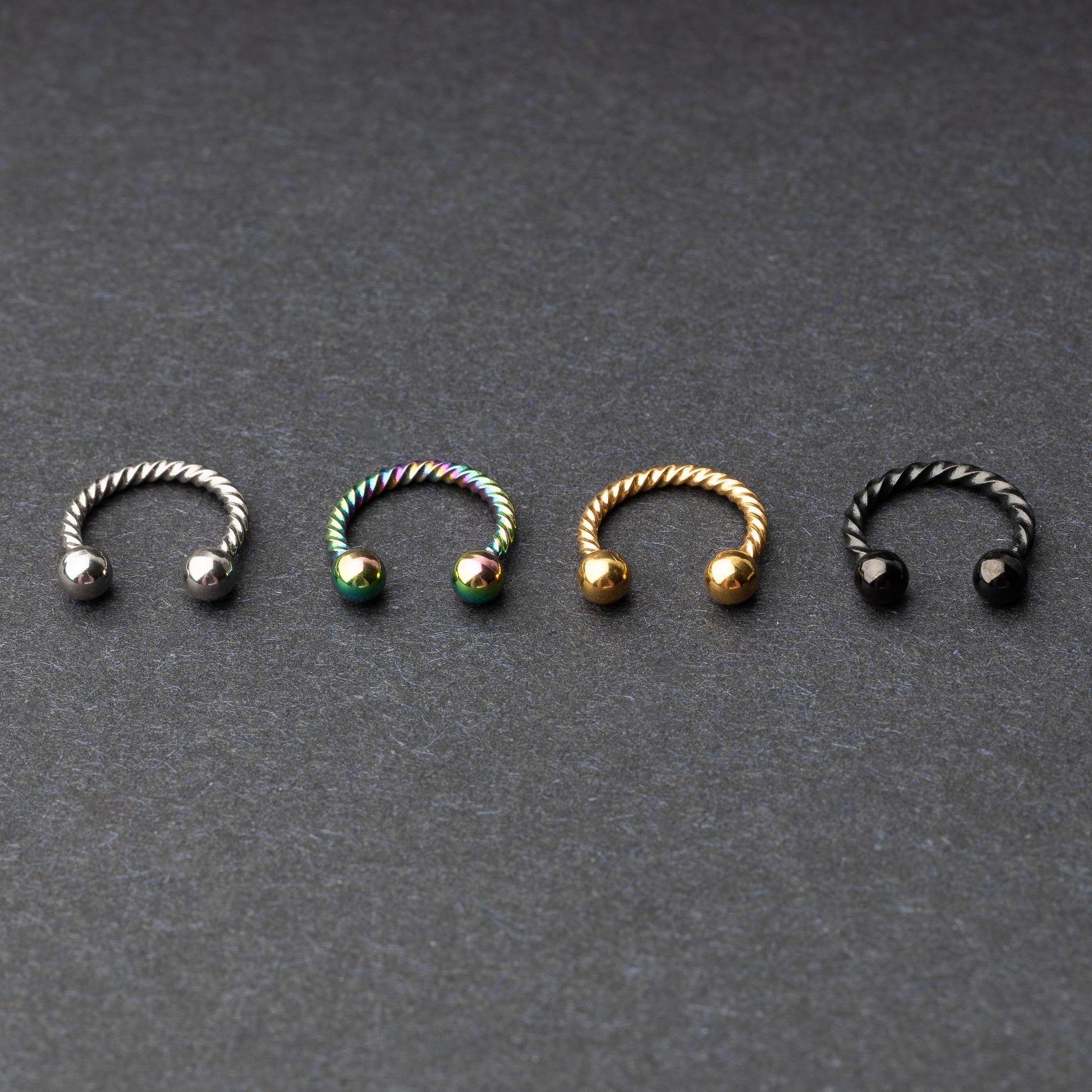 Twisted horseshoe barbells in silver, rainbow, gold, and black finishes displayed on a dark background, highlighting their bold and unique design.