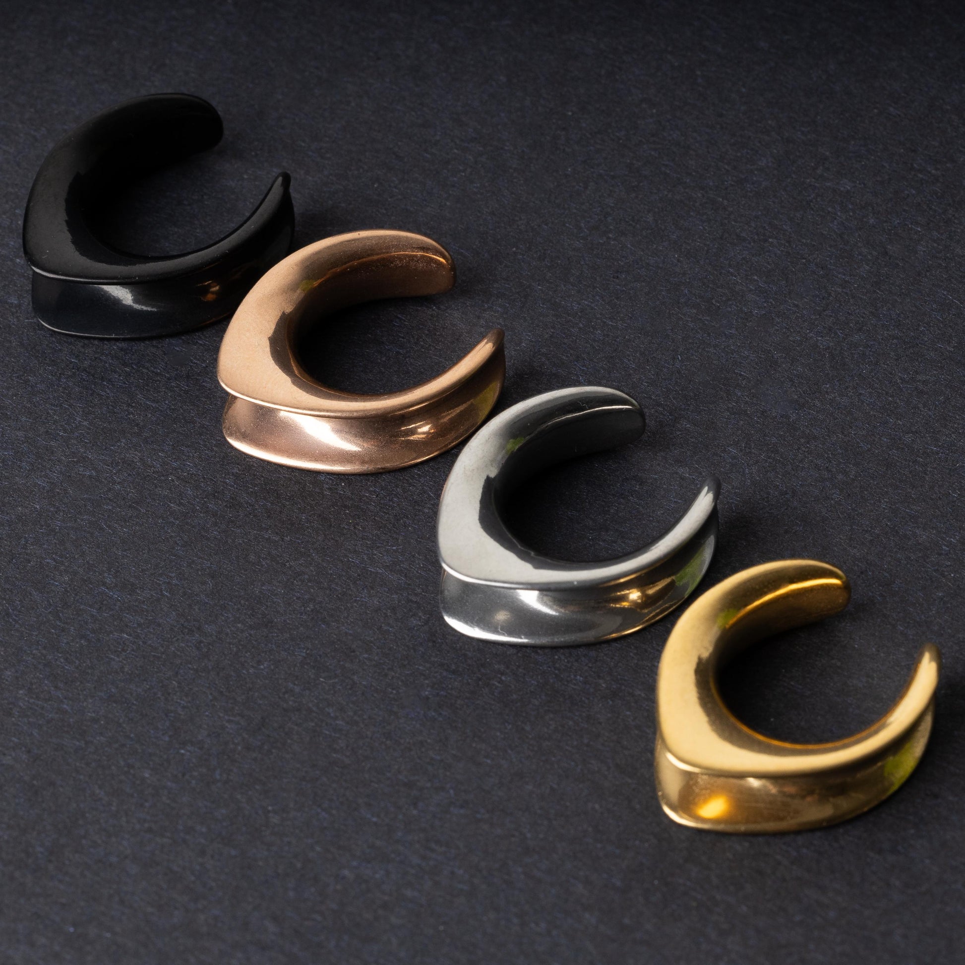 A collection of double-flared metal tunnels for stretched ears in black, gold, silver, and rose gold finishes, displayed on a dark background.
