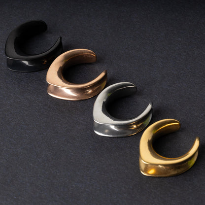 A collection of double-flared metal tunnels for stretched ears in black, gold, silver, and rose gold finishes, displayed on a dark background.
