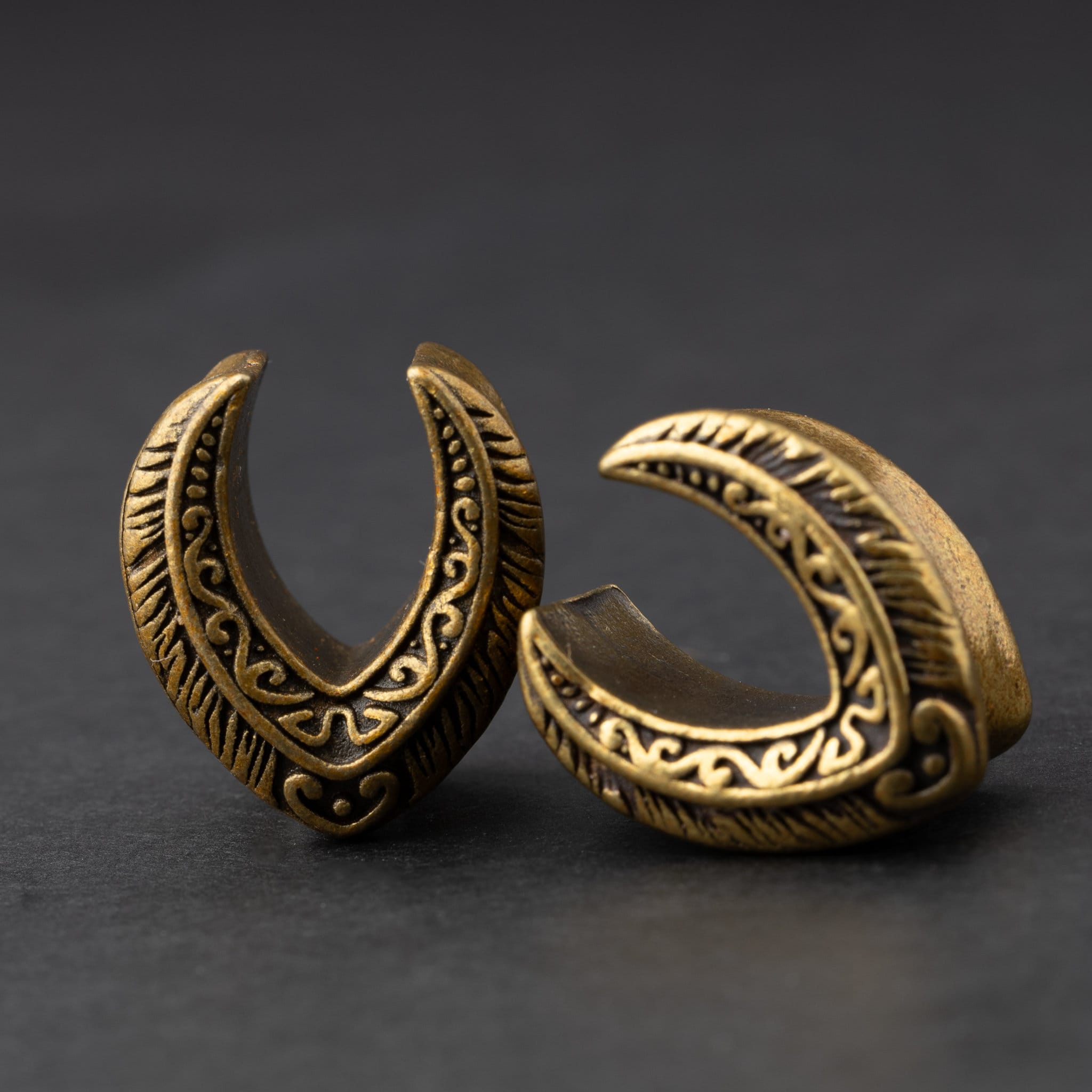 Two crescent-shaped metal tunnels for stretched ears, engraved with intricate feather and scroll designs, in a silver finish, displayed on a dark background.
