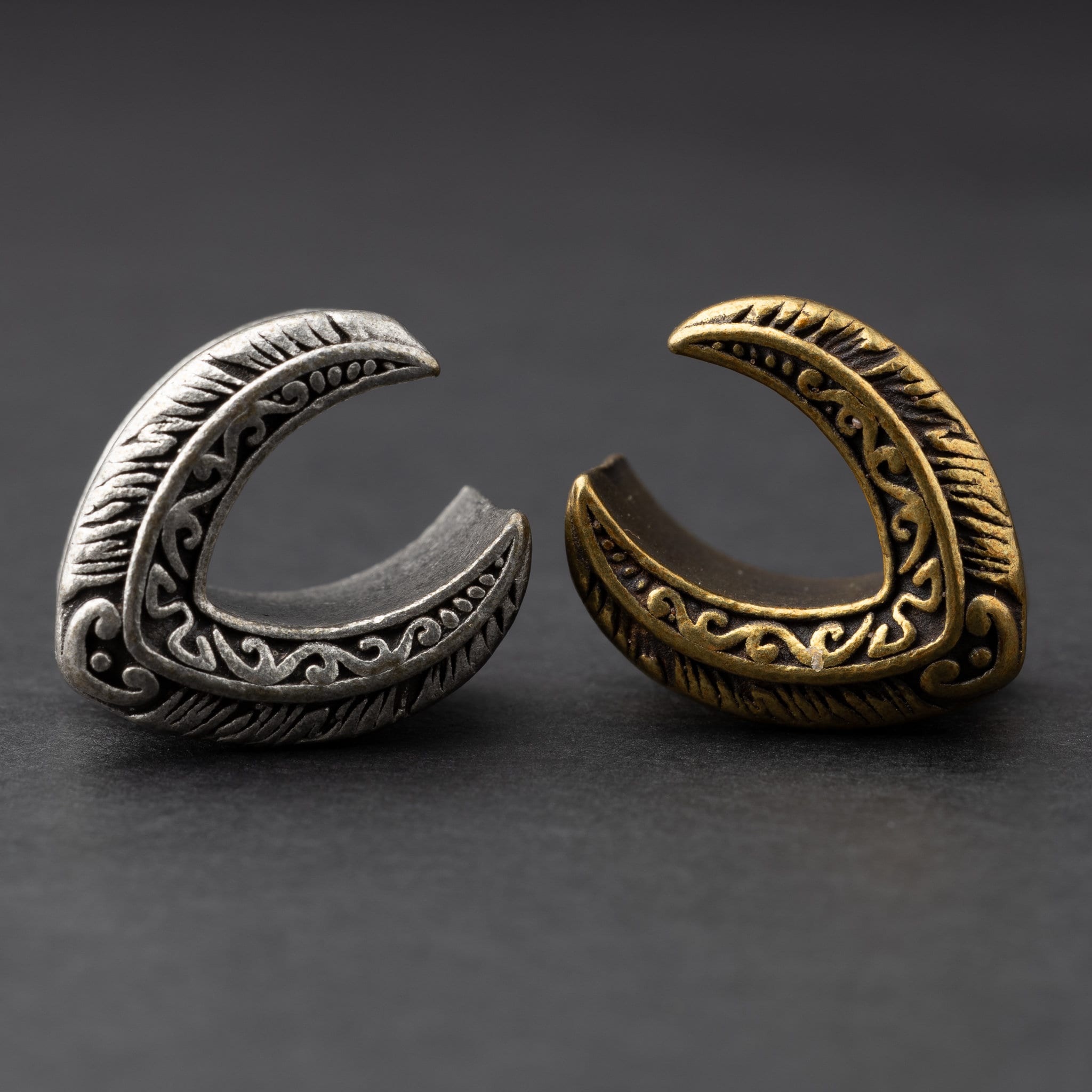 Two crescent-shaped metal tunnels for stretched ears, engraved with intricate feather and scroll designs, in silver and bronze finishes, displayed on a dark background.


