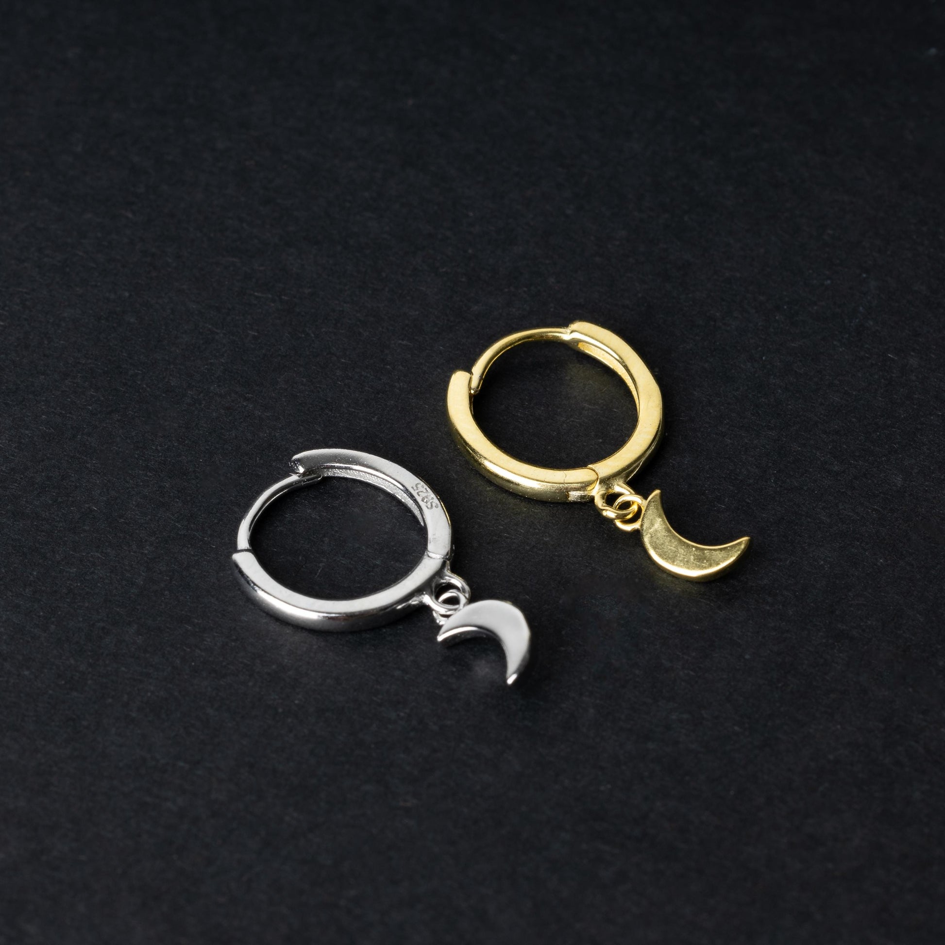 Two crescent moon huggie hoop earrings in gold and silver, each featuring a dangling crescent charm, displayed on a dark background.