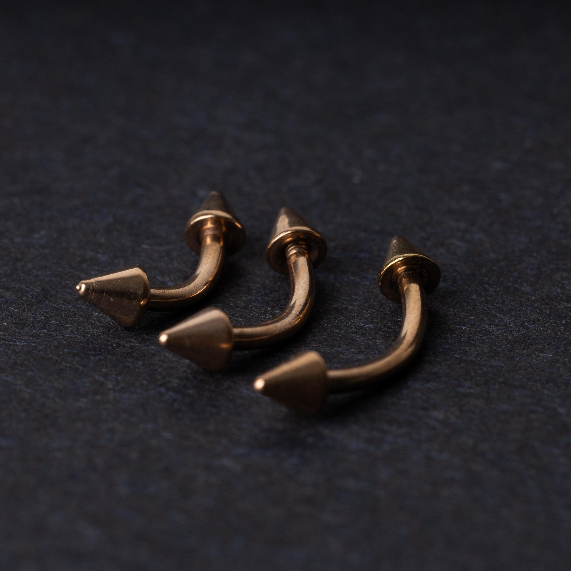 Three gold-tone titanium curved barbells with cone ends, displayed on a dark textured background, showcasing their sleek curved design and polished finish.