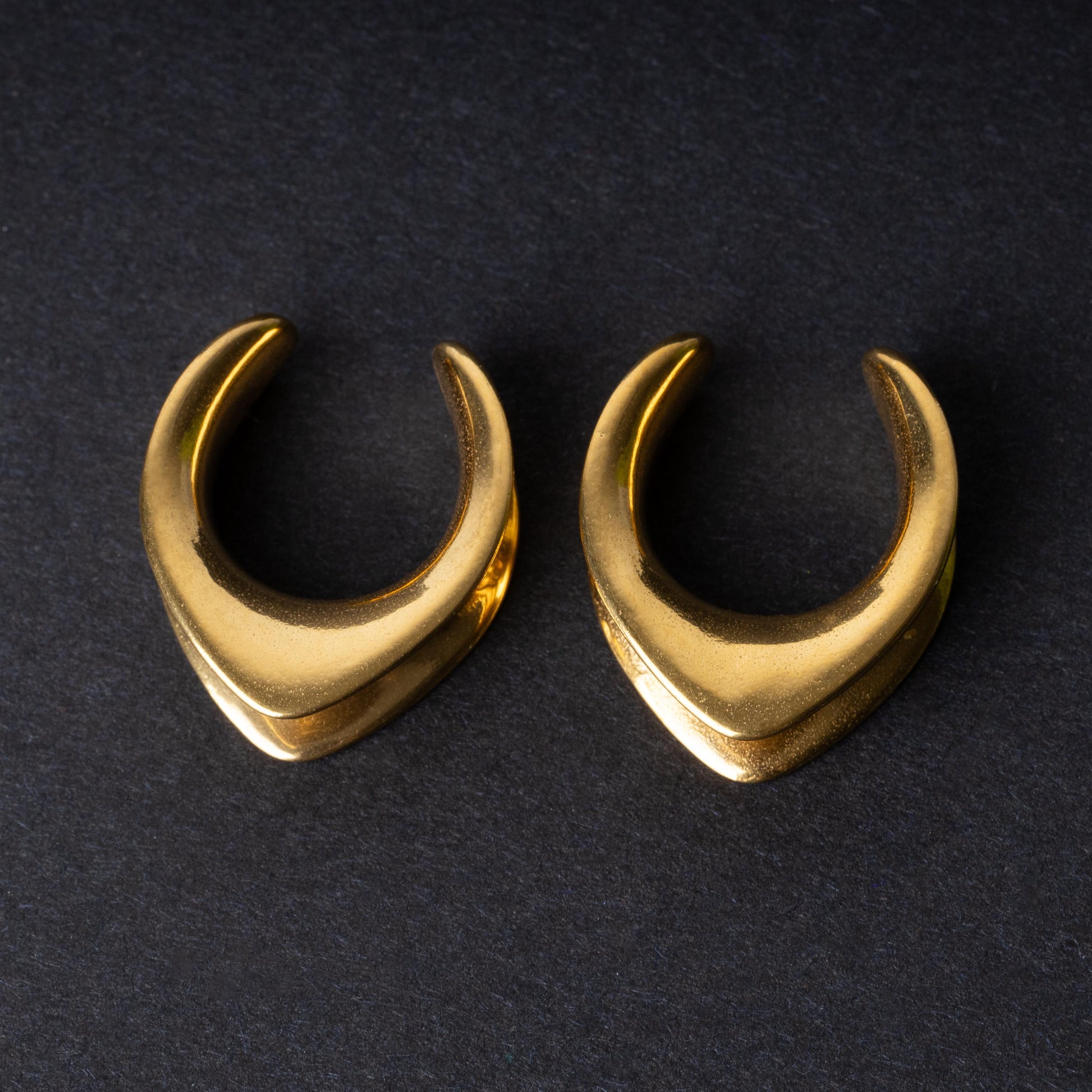 Double-flared metal tunnels for stretched ears in a gold finish, displayed on a dark background.

