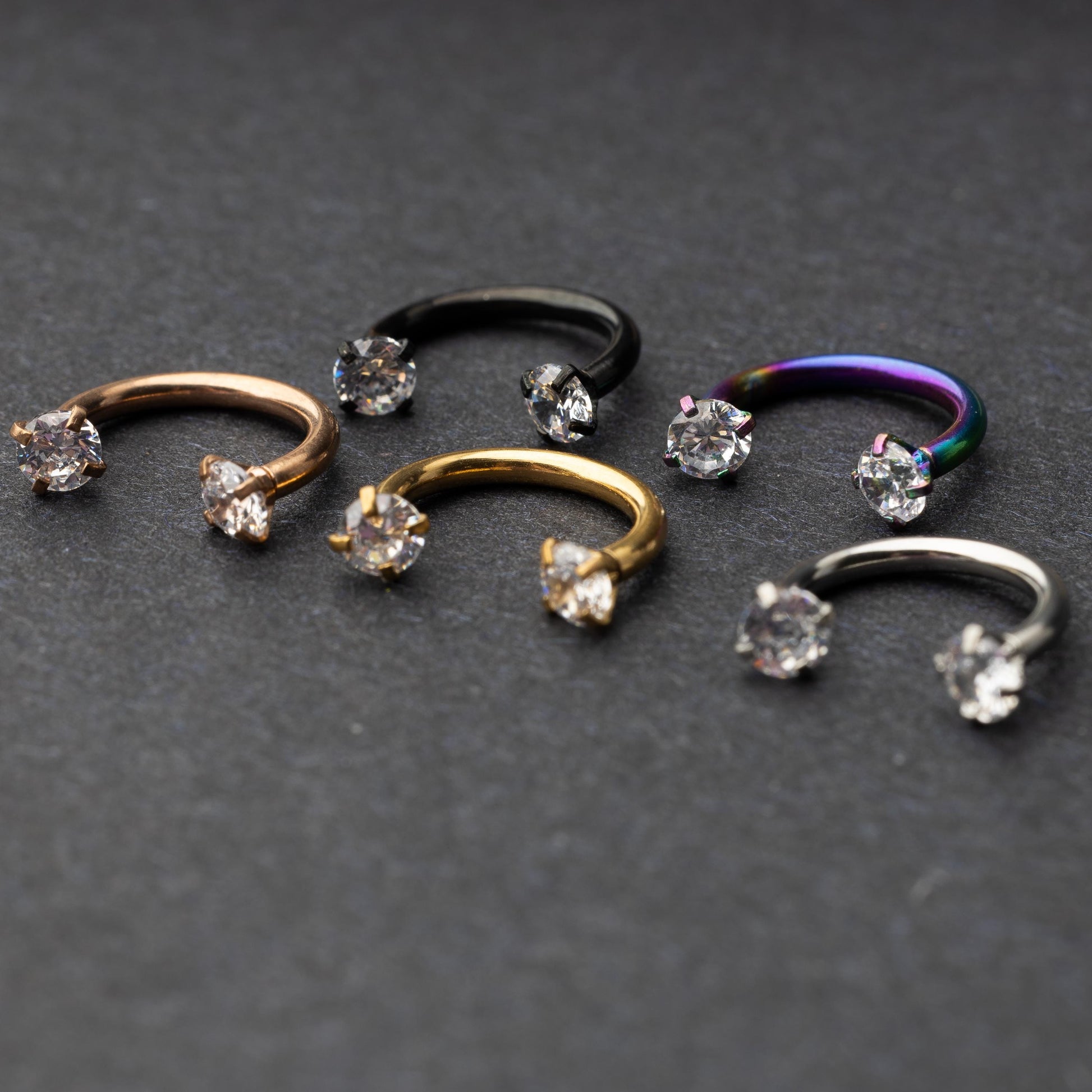 A set of five horseshoe barbells in finishes including rose gold, black, gold, rainbow, and silver, each with gemstone ends, displayed on a dark background.