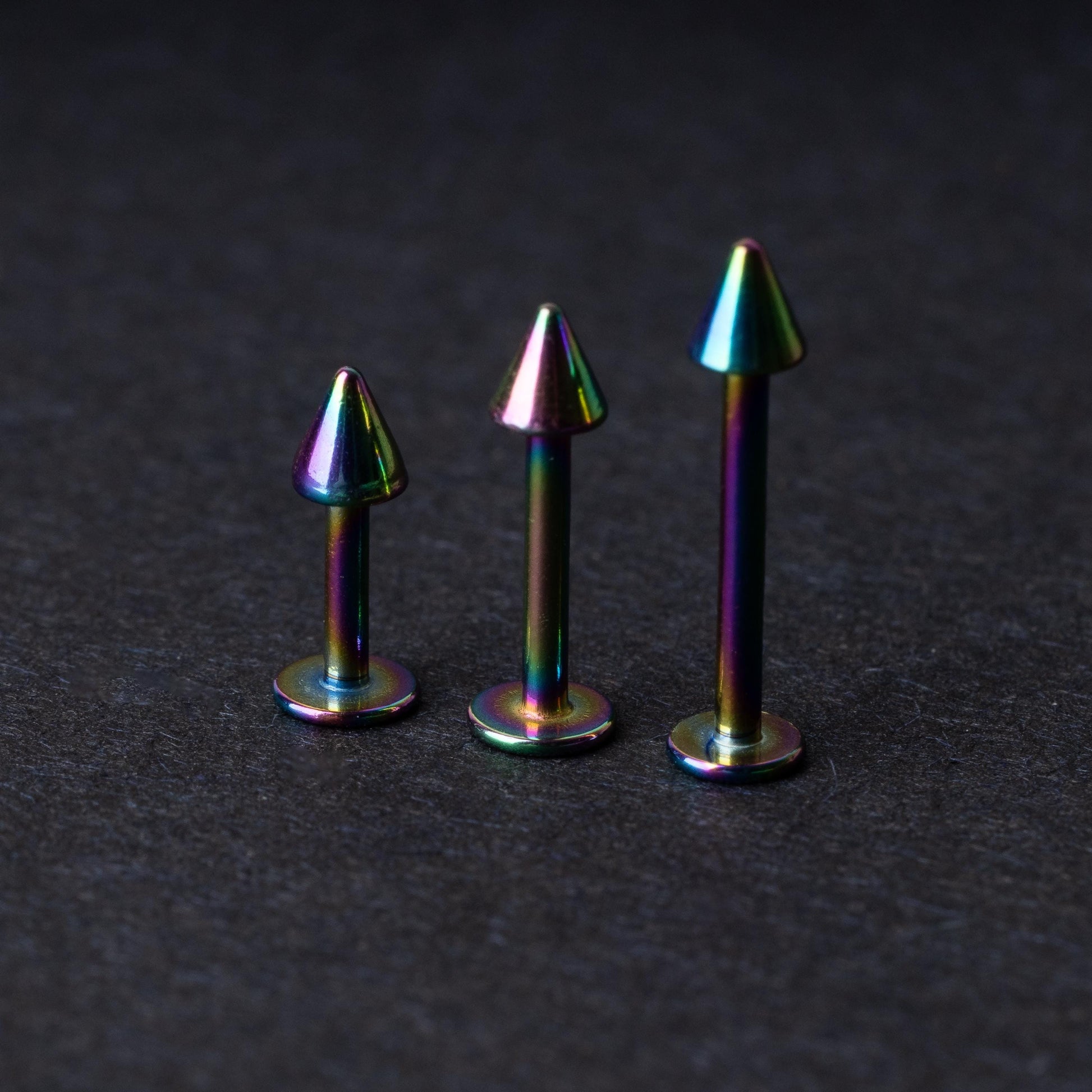 Three rainbow labret studs with cone-shaped tops displayed on a dark background, highlighting their iridescent finish and sleek, durable design.