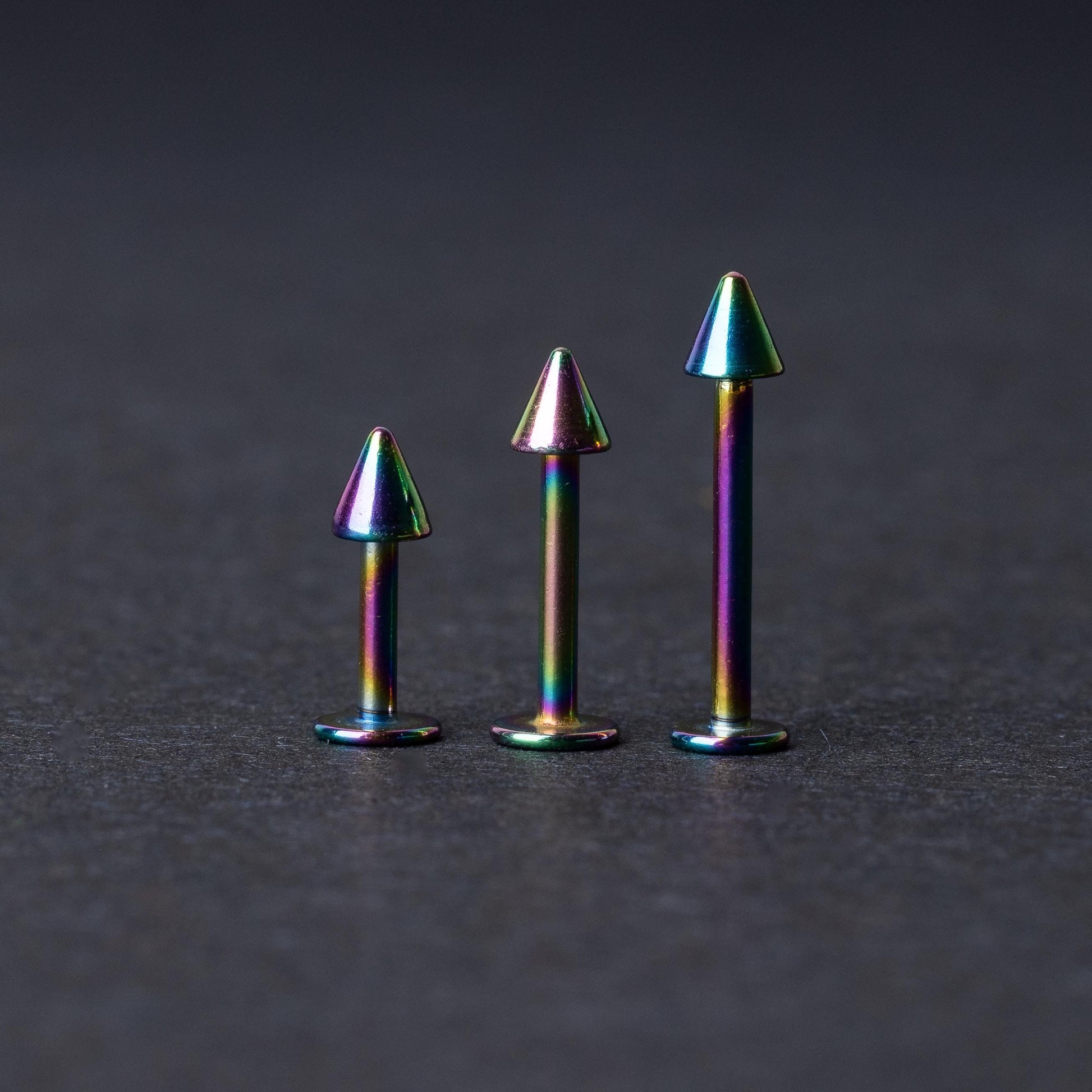 Three rainbow labret studs with cone-shaped tops displayed on a dark background, highlighting their iridescent finish and sleek, durable design.