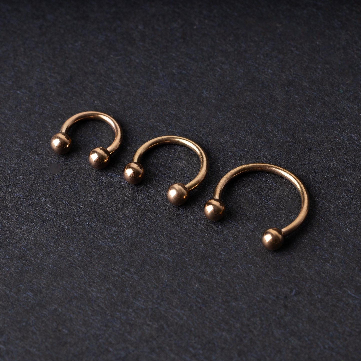 Three rose gold-tone stainless steel horseshoe hoops in varying sizes with screw-on ball ends, displayed against a dark background.