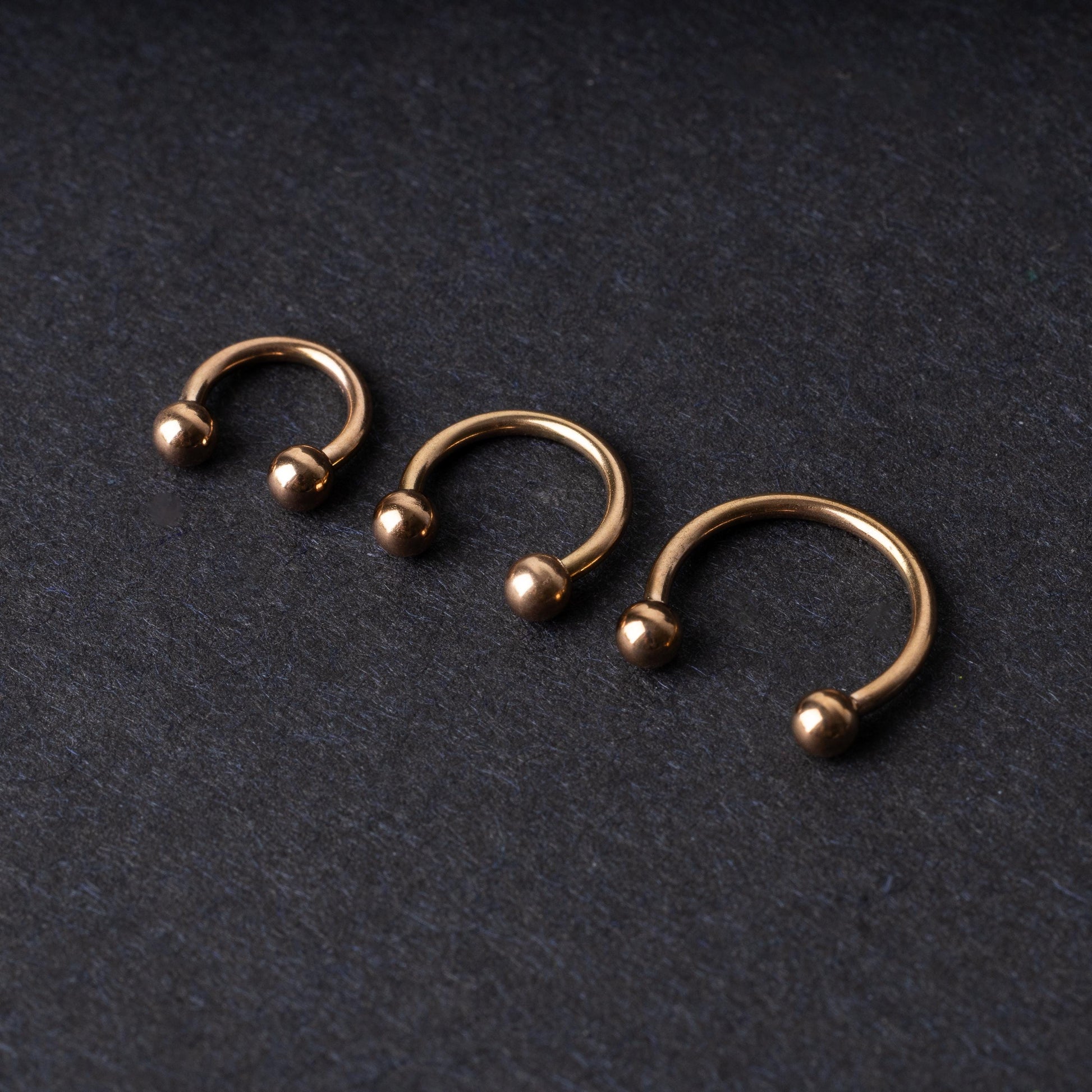 Three rose gold-tone stainless steel horseshoe hoops in varying sizes with screw-on ball ends, displayed against a dark background.