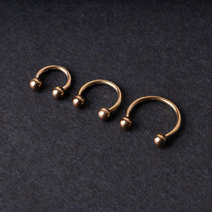 Three rose gold-tone stainless steel horseshoe hoops in varying sizes with screw-on ball ends, displayed against a dark background.