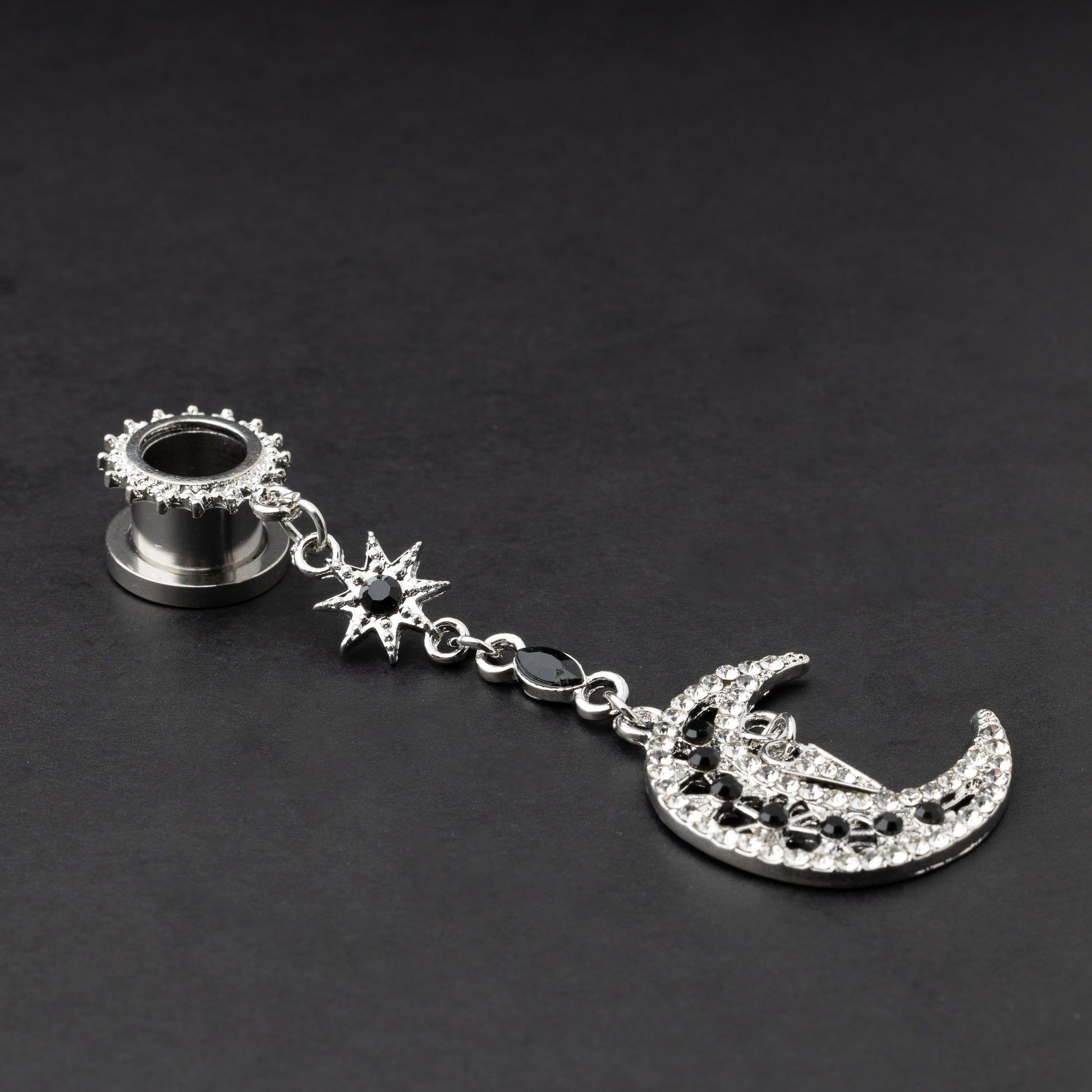 A pair of silver ear hangers for stretched ears with dangling crescent moon and star designs, adorned with black and clear rhinestones, displayed on a dark background.