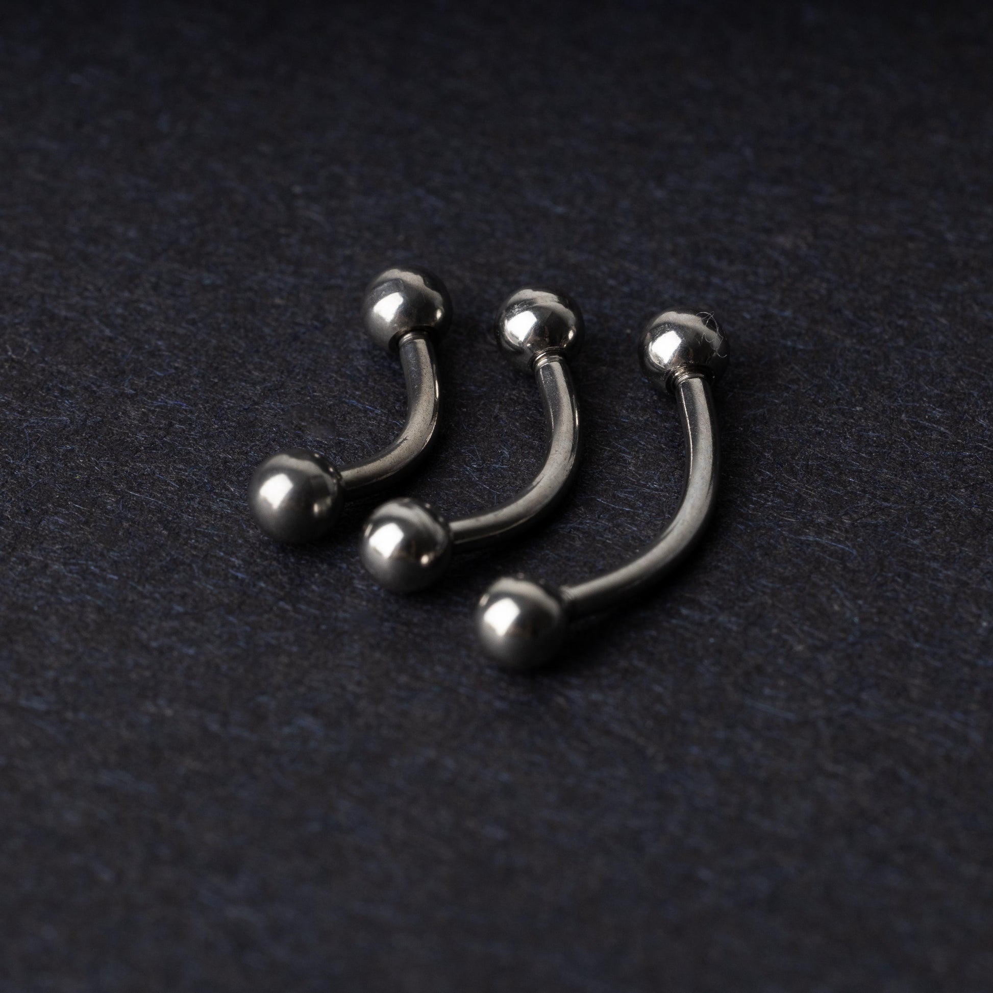 Three silver stainless steel curved barbells with polished ball ends, displayed on a dark textured background.