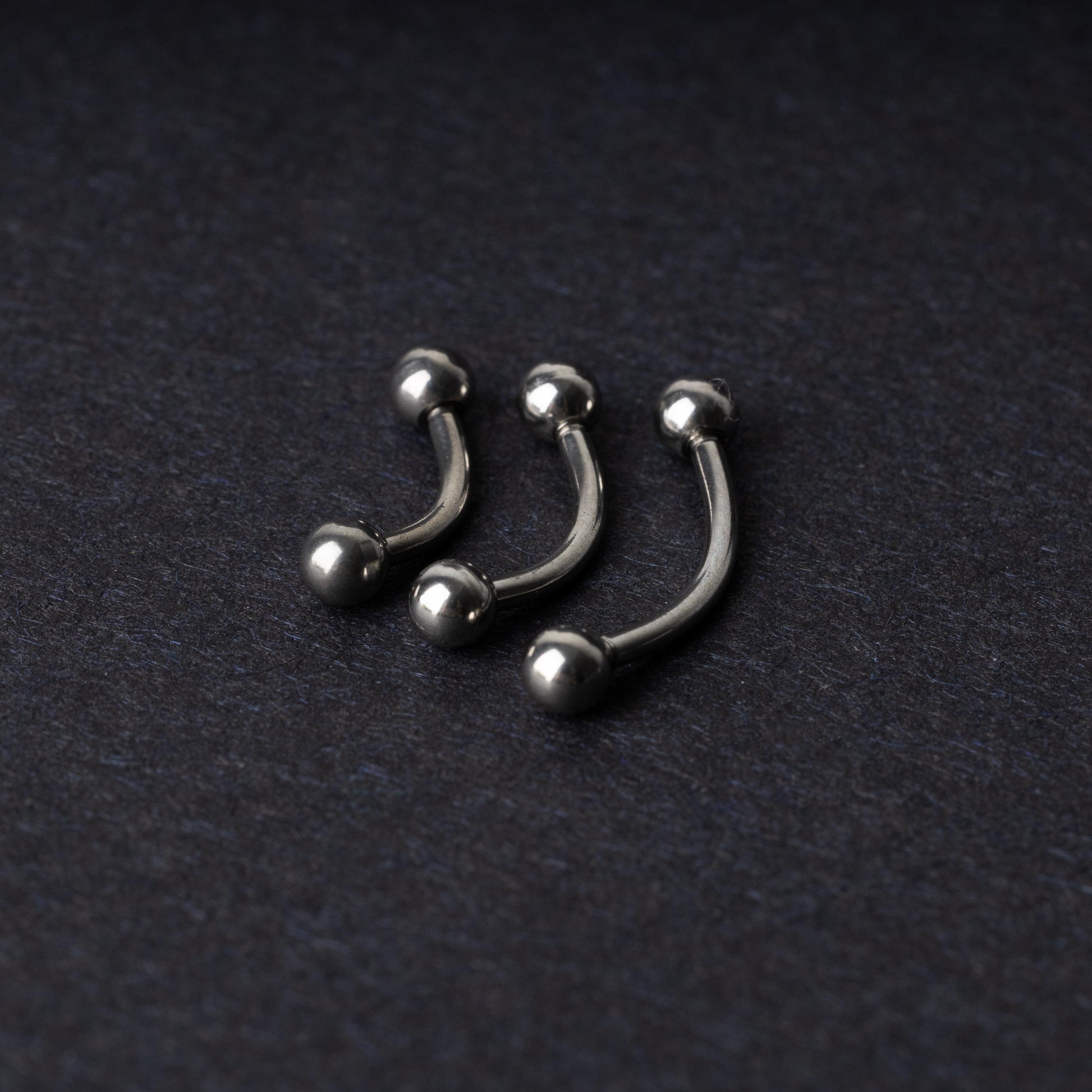 Three silver stainless steel curved barbells with polished ball ends, displayed on a dark textured background.
