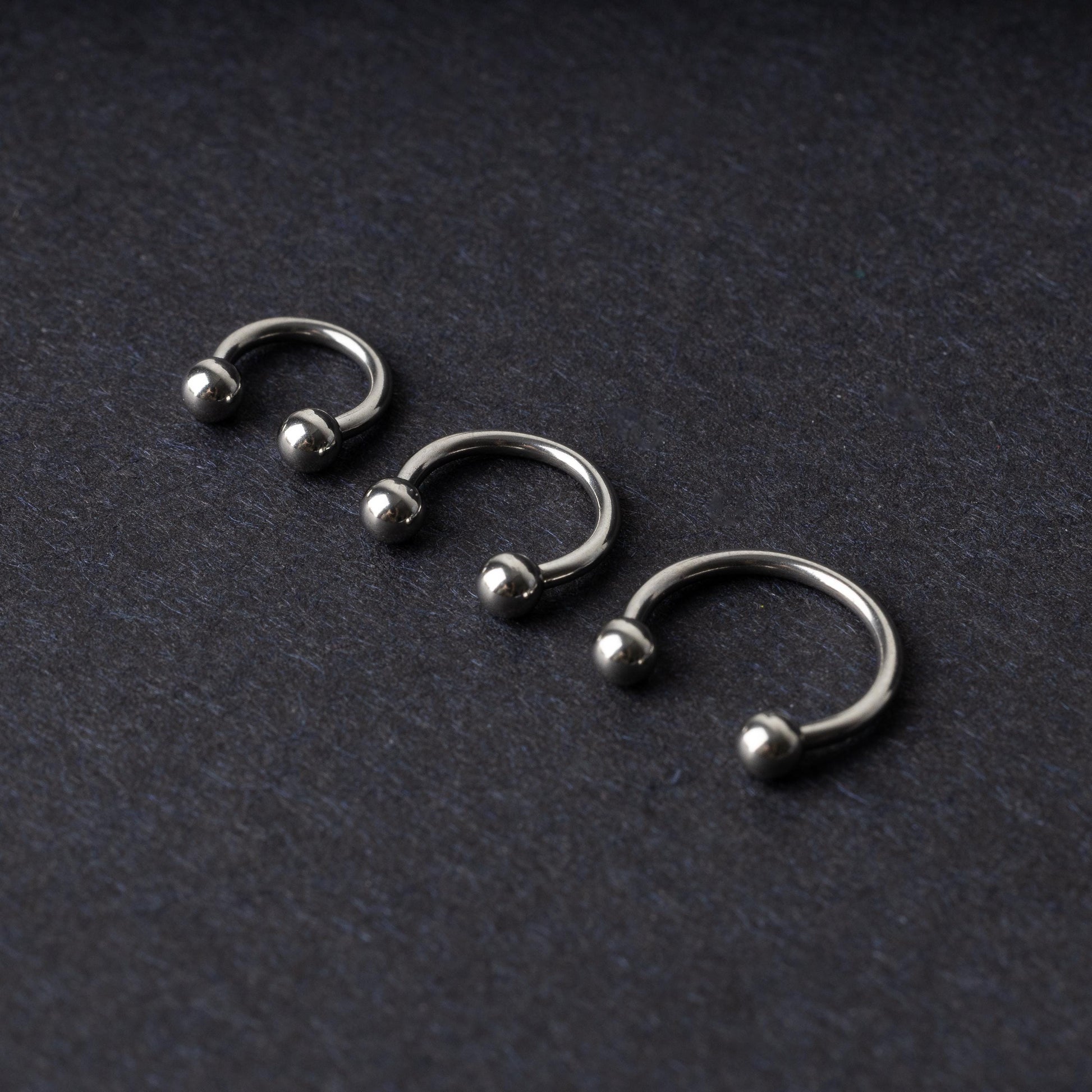 Three silver stainless steel horseshoe hoops with polished ball ends, displayed on a dark textured background.