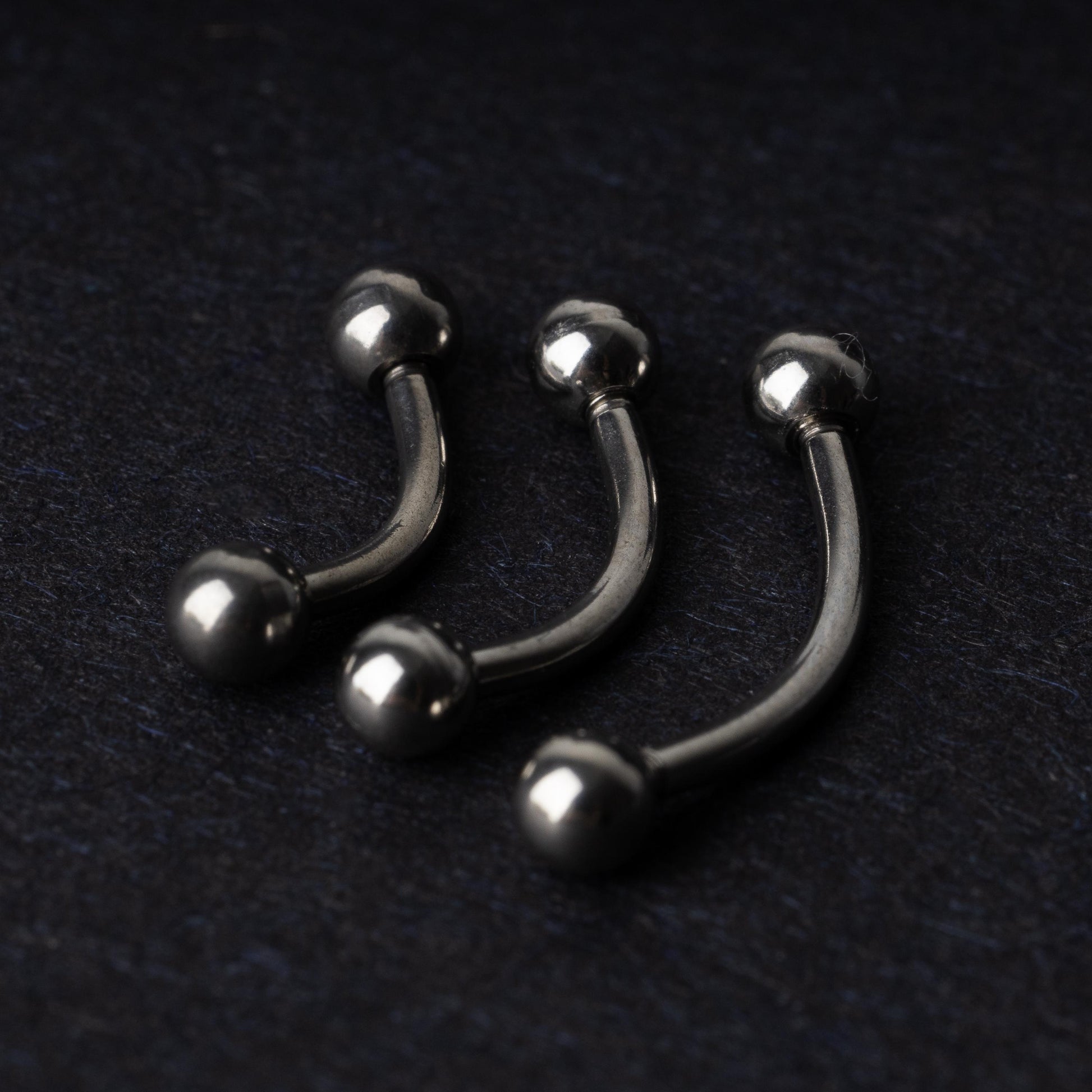 Three silver stainless steel curved barbells with polished ball ends, displayed on a dark textured background.