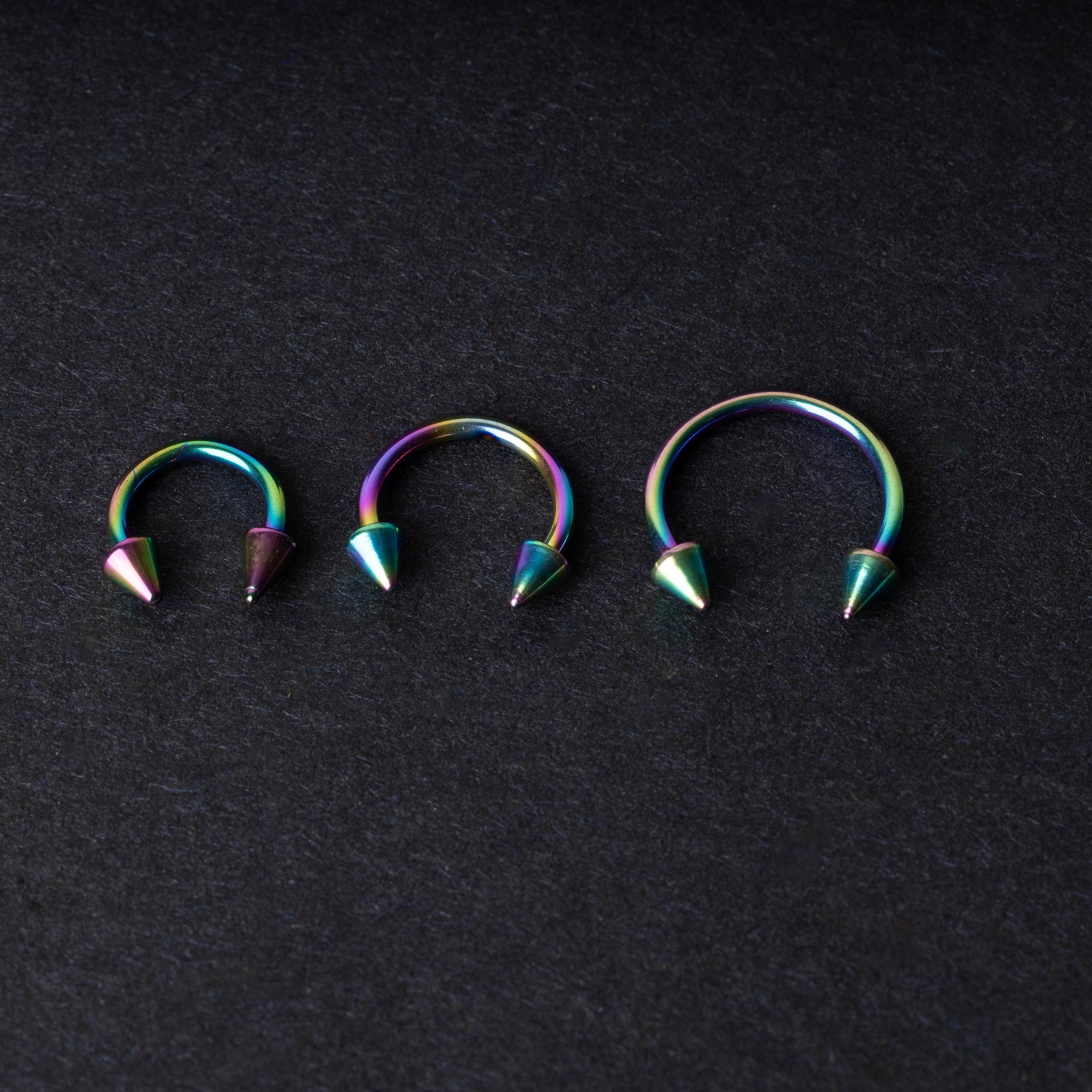 Three stainless steel horseshoe hoops with cone-shaped ends and a rainbow anodized finish displayed on a dark background, showcasing their bold design and vibrant colors.