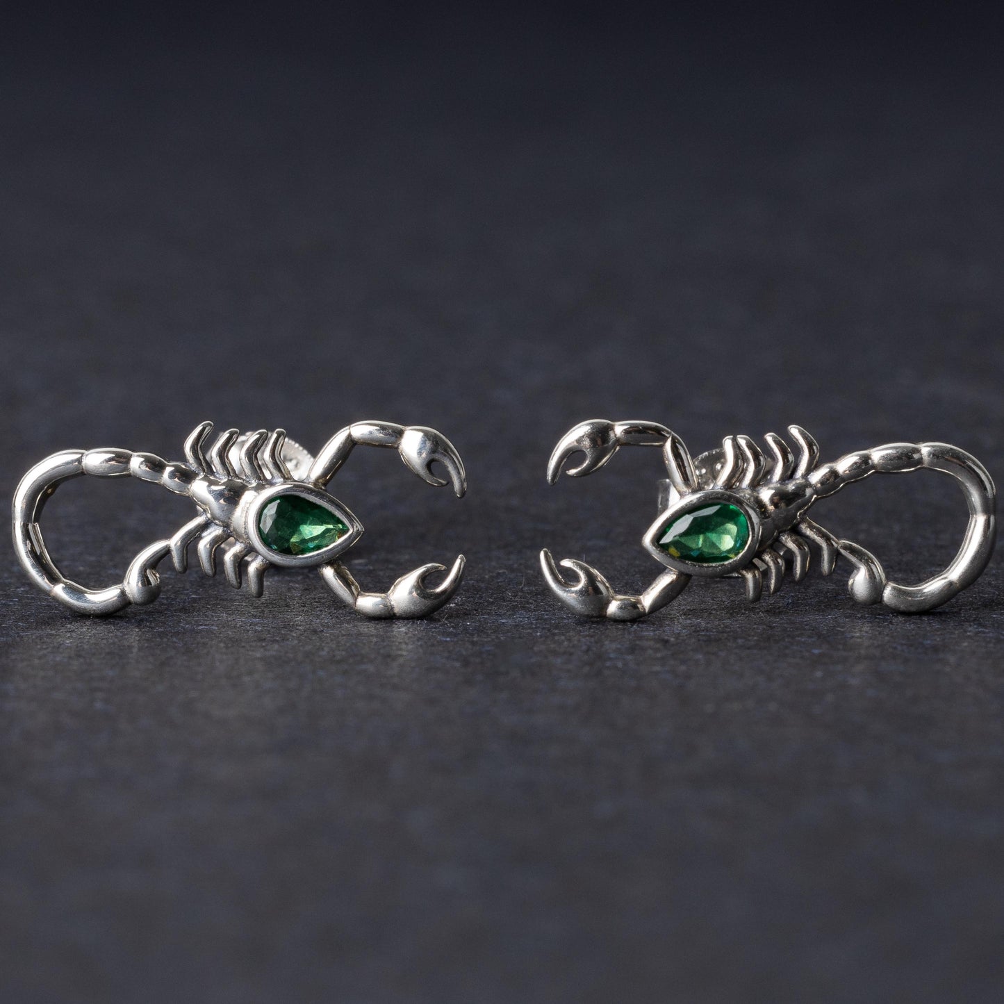A pair of 925 sterling silver scorpion-shaped earrings with green gemstone accents as eyes, displayed on a dark background.