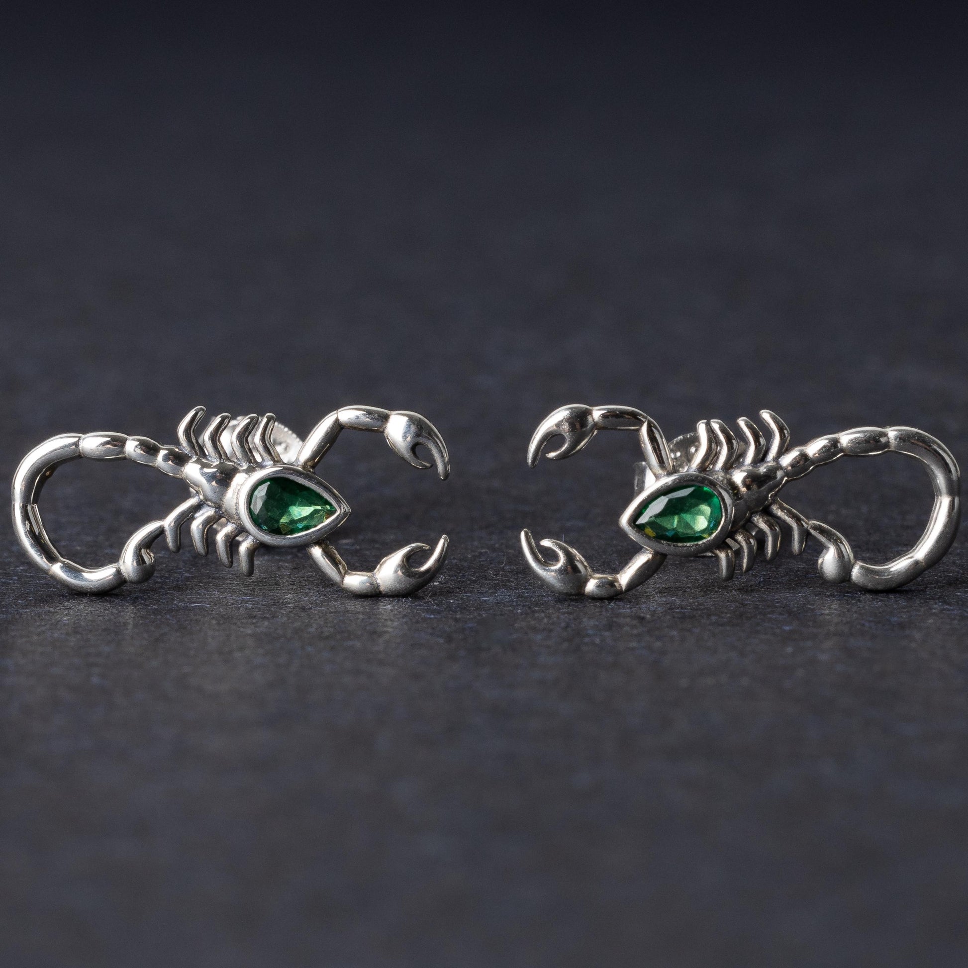 A pair of 925 sterling silver scorpion-shaped earrings with green gemstone accents as eyes, displayed on a dark background.