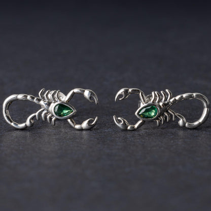 A pair of 925 sterling silver scorpion-shaped earrings with green gemstone accents as eyes, displayed on a dark background.