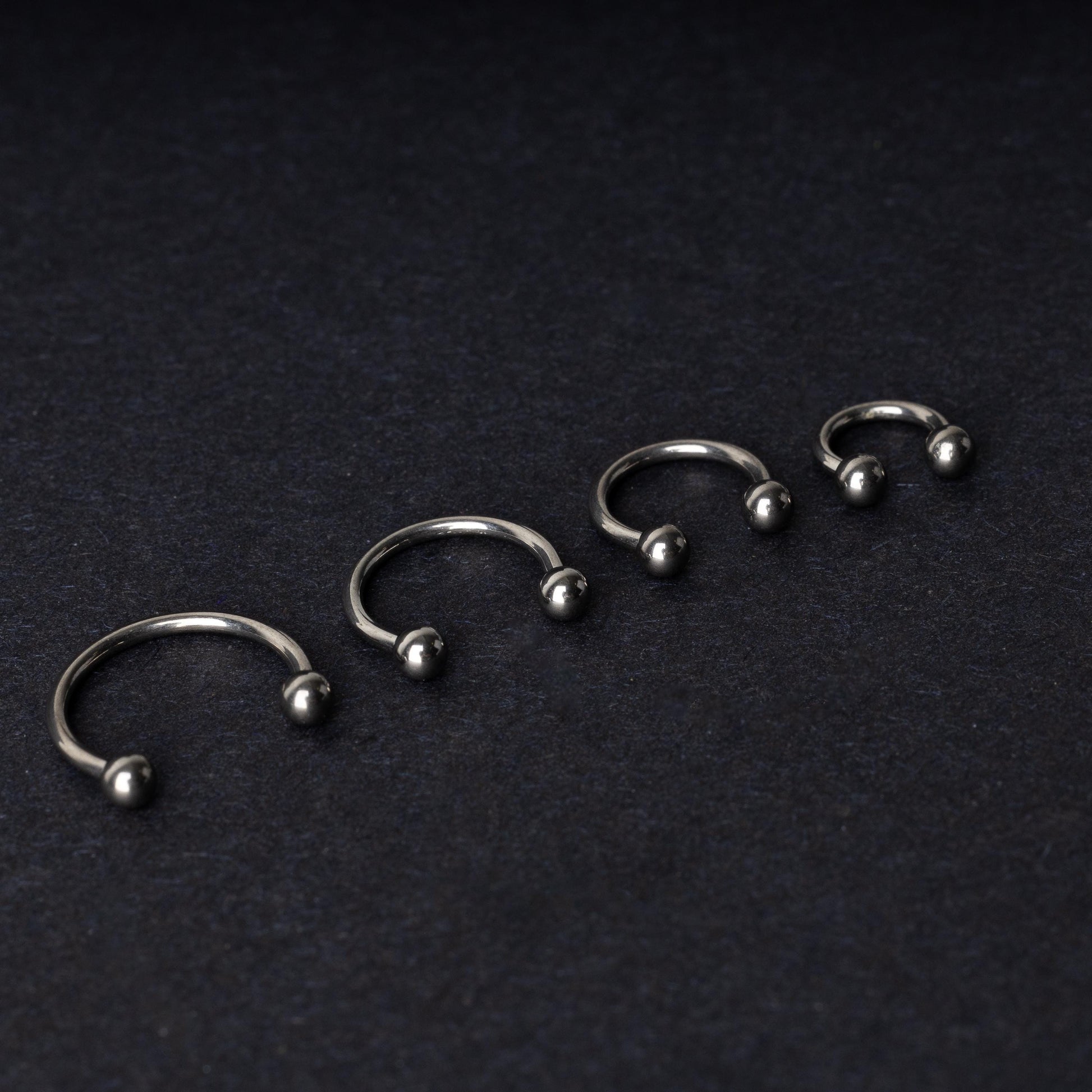 Four titanium horseshoe hoops with ball ends, arranged from largest to smallest on a dark background.