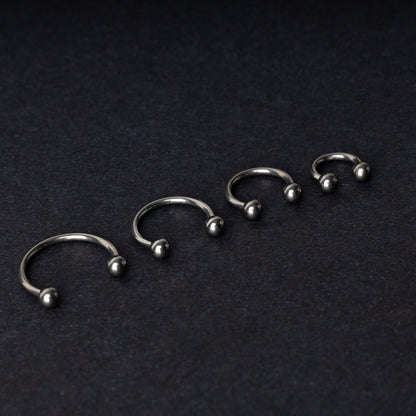 Four titanium horseshoe hoops with ball ends, arranged from largest to smallest on a dark background.