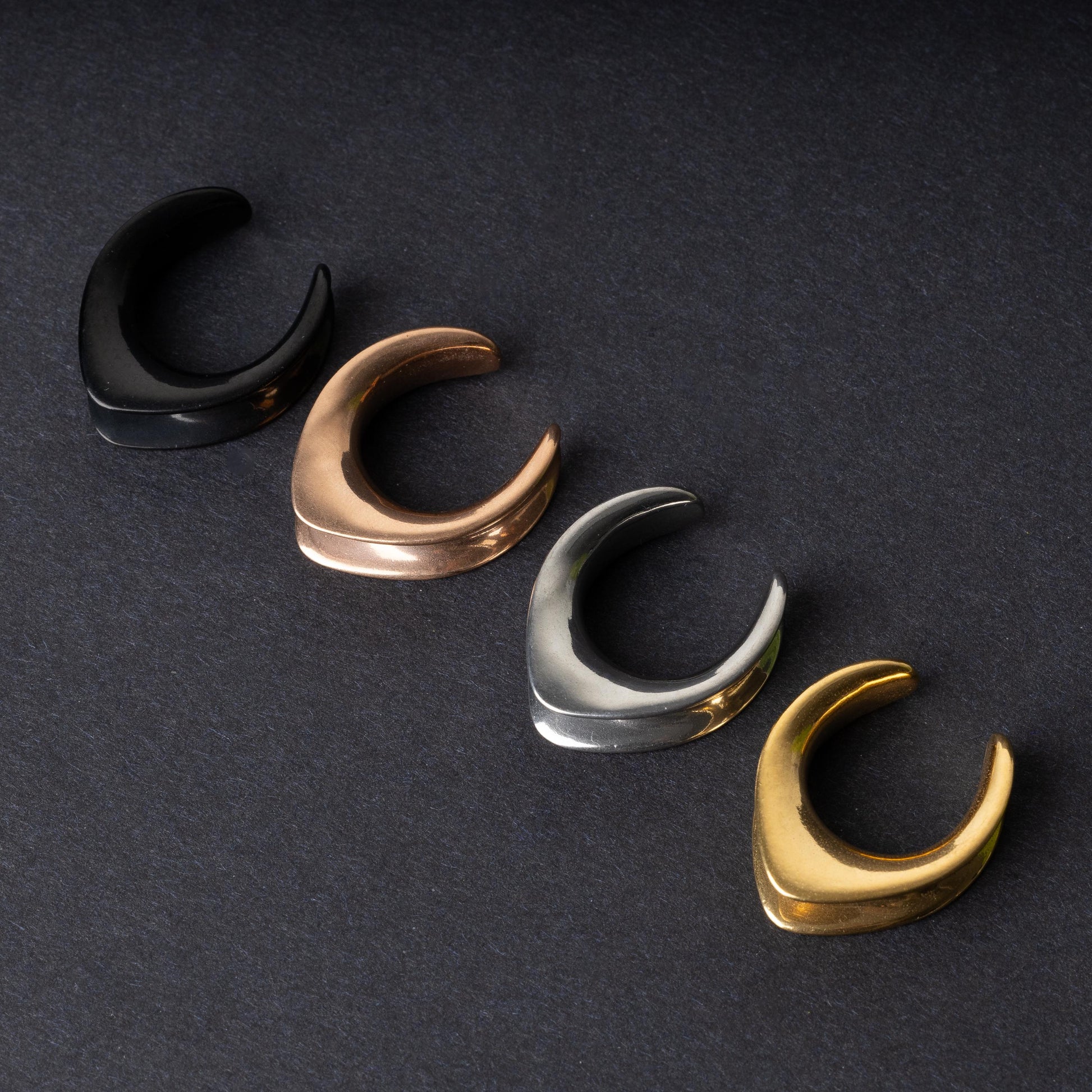 A collection of double-flared metal tunnels for stretched ears in black, gold, silver, and rose gold finishes, displayed on a dark background.

