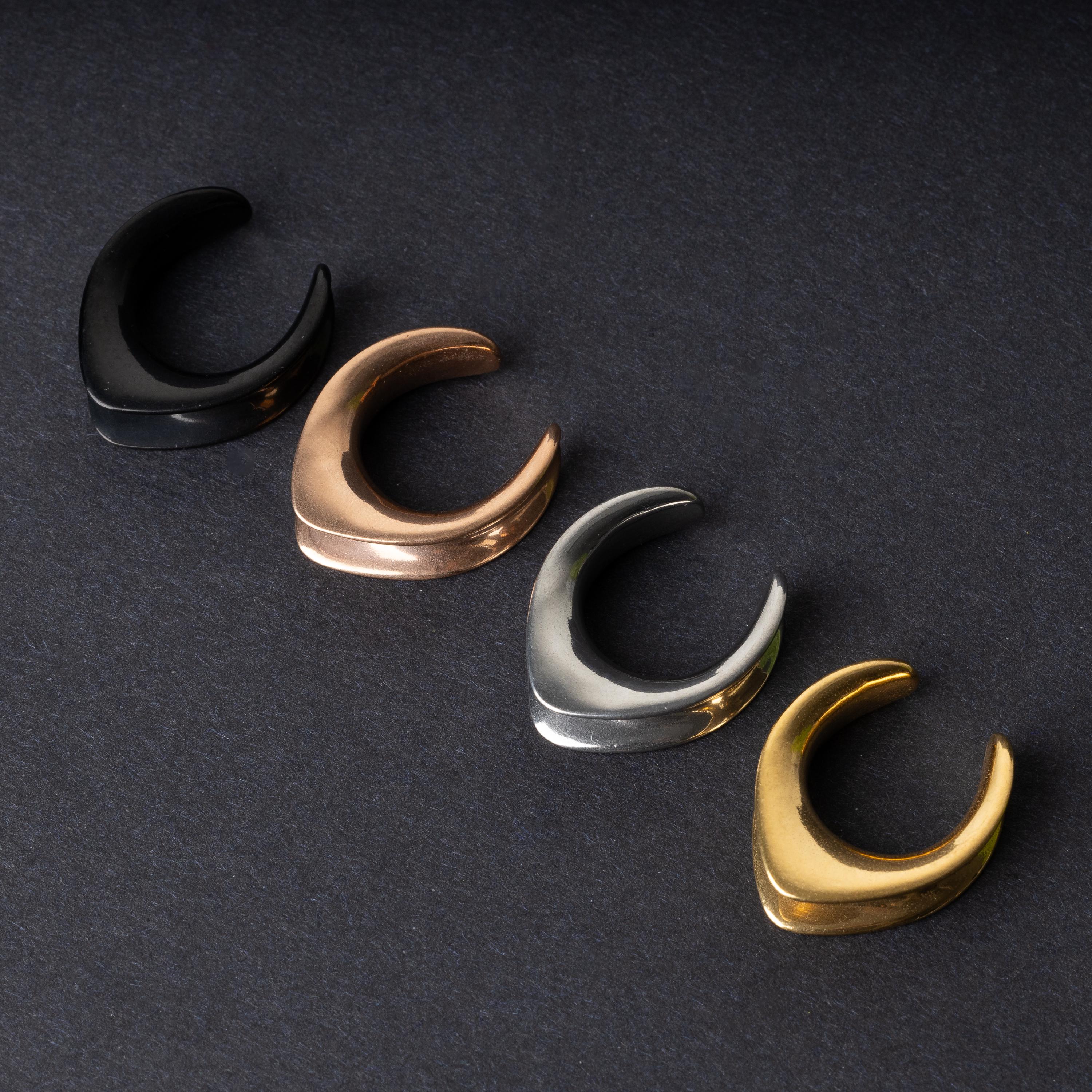 A collection of double-flared metal tunnels for stretched ears in black, gold, silver, and rose gold finishes, displayed on a dark background.

